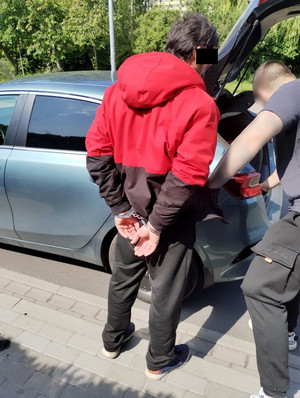 Zatrzymany obywatel Mołdawii w kajdankach na rękach i policjant przed autem.