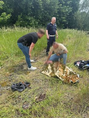 Dwaj mężczyźni przy leżącym na ziemi odnalezionym mężczyzną, owiniętym w koc termiczny. Z tyłu stoi umundurowany policjant.