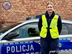 Policjantka w mundurze i nałożonej na niego żółtej kamizelce z napisem Policja stoi przed radiowozem.