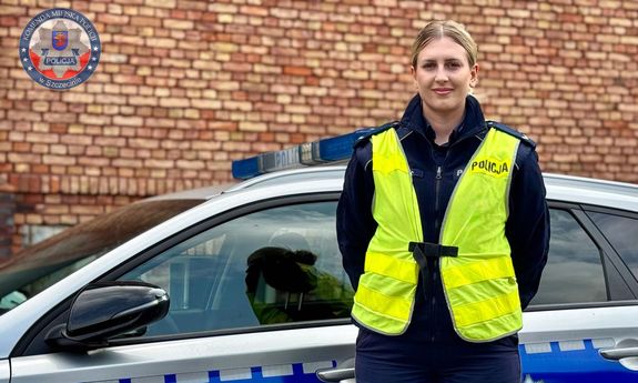 Policjantka w mundurze i nałożonej na niego żółtej kamizelce z napisem Policja stoi przed radiowozem.