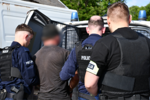 Trzej policjanci z zatrzymanym mężczyzną przy radiowozie.