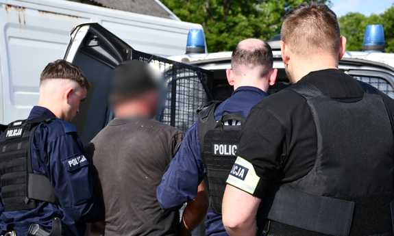 Trzej policjanci z zatrzymanym mężczyzną przy radiowozie.