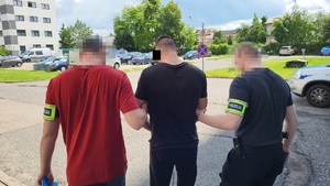 Dwaj funkcjonariusze prowadzą zakutego w kajdanki mężczyznę.