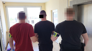 Dwaj funkcjonariusze prowadzą zakutego w kajdanki mężczyznę.
