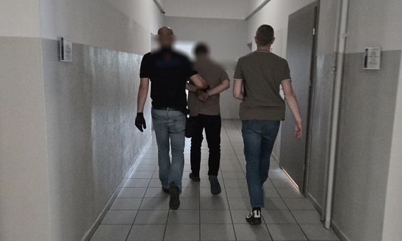 Dwaj policjanci w cywilnych ubraniach prowadzą skutego w kajdanki mężczyznę korytarzami komendy.