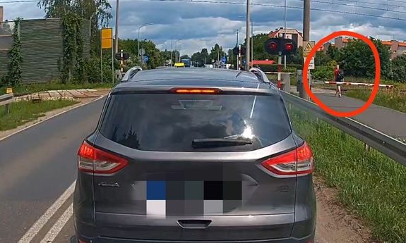 Pieszy przechodzi przez przejazd kolejowy na czerwonym świetle i przy opuszczonych rogatkach.