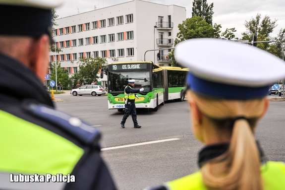 Dwoje policjantów w białych czapkach obserwuje funkcjonariusza kierującego ruchem na skrzyżowaniu.