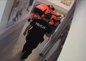 Ratownicy pochyleni nad odnalezionym mężczyzną. Na pierwszym planie widoczny idący do nich korytarzem policjant.