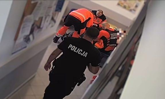 Ratownicy pochyleni nad odnalezionym mężczyzną. Na pierwszym planie widoczny idący do nich korytarzem policjant.