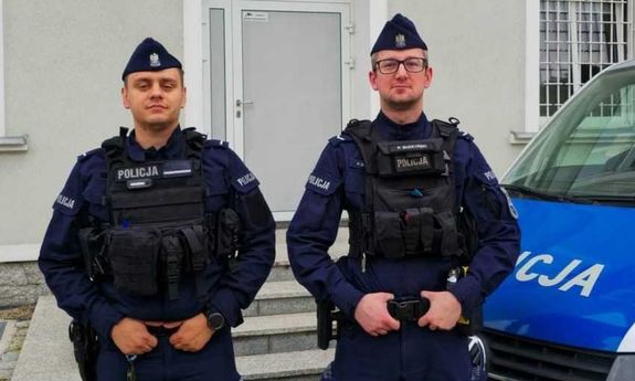 dwóch policjantów stoi obok radiowozu i pozują do zdjęcia.