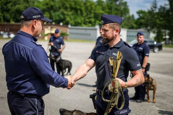 Policjant ściska dłoń drugiemu policjantowi, który w drugiej ręce trzyma puchar. Z tyłu widać przewodników z psami służbowymi.