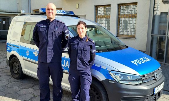 Dwoje umundurowanych policjantów przed radiowozem.
