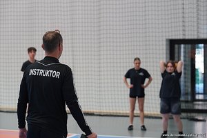 Na zdjęciu mężczyzna z koszulką z napisem INSTRUKTOR obserwujący kobietę trzymającą piłkę za głową, która będzie nią rzucać. W tle kobieta i mężczyzna obserwujący kobietę z piłką.