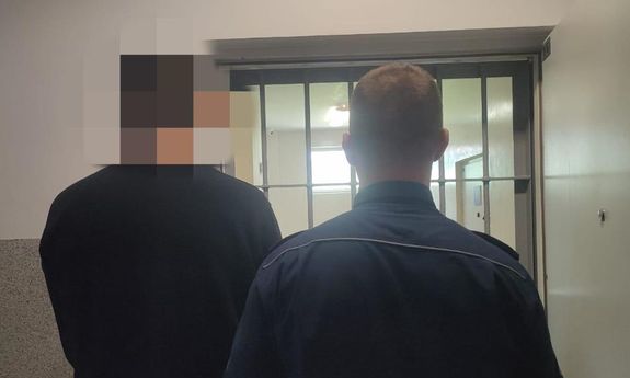 Zatrzymany mężczyzna prowadzony przez policjanta - z tyłu widoczne są kraty.