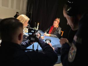 Na zdjęciu dwie kobiety siedzące przy stole. Przed nimi stoją dwaj umundurowani policjanci, którzy trzymając kamerę i mikrofon nagrywają je. W tle stoi kolejna kobieta.