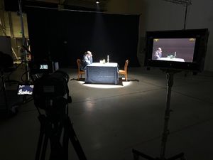 Miejsce nagrywania materiału filmowego. Przy stole siedzi kobieta, na którą skierowana jest kamera, a obraz z tej kamery widoczny jest na monitorze znajdującym się na statywie stojącym obok kamery.