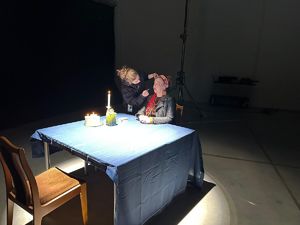 Przy stole siedzi kobieta, a obok niej stoi druga, która ją maluje