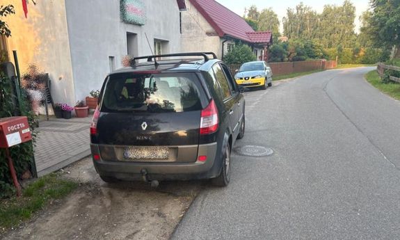 Na zdjęciu ulica, a przy niej zaparkowane dwa samochody i budynki stojące obok drogi