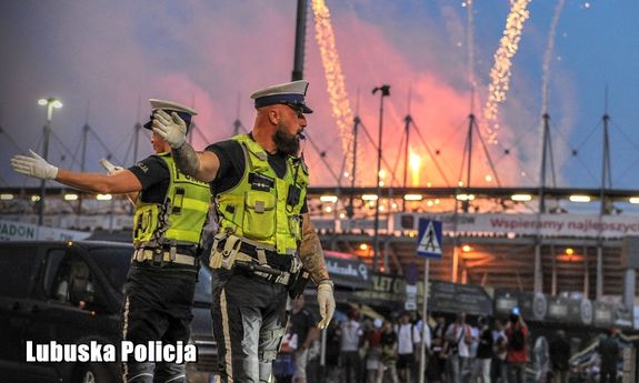 Na zdjęciu dwóch umundurowanych policjantów ruchu drogowego, a za nimi w tle stadion.
