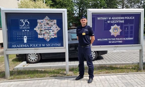 Policjant stoi przy tablicach z napisem Akademia Policji w Szczytnie.