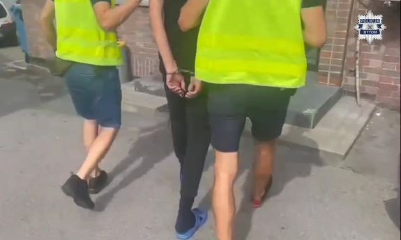 Dwaj policjanci w żółtych kamizelkach prowadzą zatrzymanego. Widok z tyłu.
