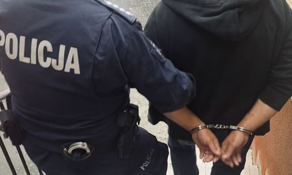 Policjant prowadzi zatrzymanego mężczyznę zakutego w kajdanki.