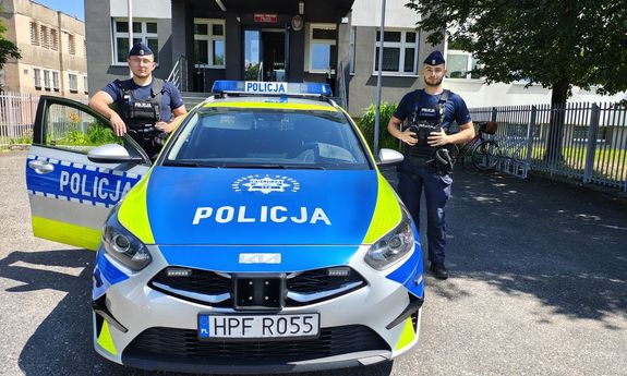 Przed budynkiem komendy z po obu stronach radiowozu oznakowanego stoją umundurowani policjanci.