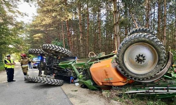Ciągnik rolniczy leżący na boku na jezdni. Z boku widać policjanta a z tyłu radiowóz.
