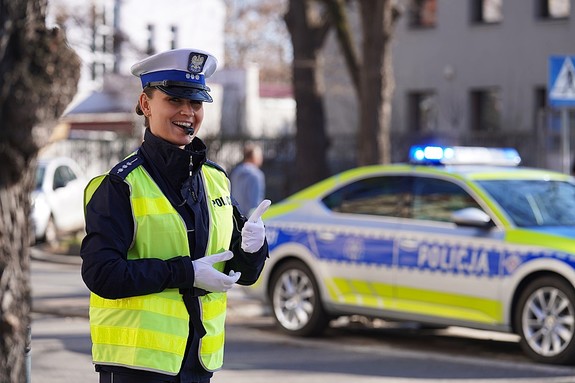 Policjantka ruchu drogowego z gwizdkiem w ustach stoi na ulicy, w oddali widać policyjny radiowóz.