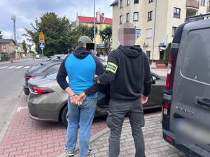 Ubrany po cywilnemu policjant, w opasce na ręce z napisem Policja, prowadzi zatrzymanego do samochodu.