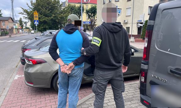 Ubrany po cywilnemu policjant, w opasce na ręce z napisem Policja, prowadzi zatrzymanego do samochodu.