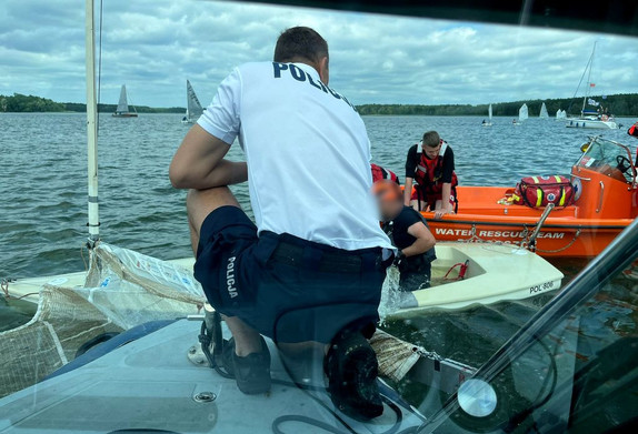 Policjant na łodzi obok w wodzie żeglarze, którym udzielono pomocy.Z tyłu widoczny ratownik WOPR.