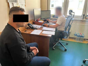 Zatrzymany w kajdankach siedzi na krześle. Z tyłu widać policjanta przy biurku.