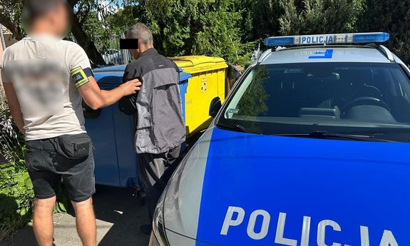 Policjant trzyma zatrzymanego mężczyznę, za nimi stoją kosze na śmieci, a obok nich oznakowany, policyjny radiowóz.