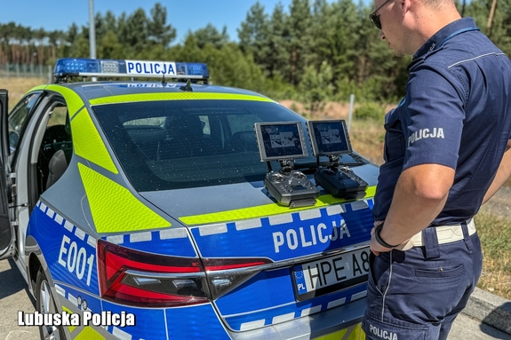 Policjant stoi za radiowozem i na klapie bagażnika ma ustawione dwie konsole do obsługi drona.