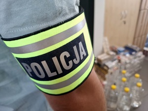 Na pierwszym planie widoczna opaska na ramieniu z napisem Policja. w tle widać zabezpieczone butelki z nielegalnym alkoholem.