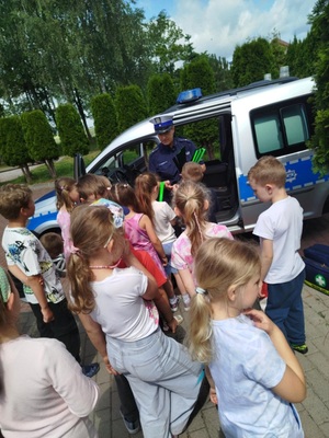 Policjantka stoi przy radiowozie i rozdaje odblaski grupie dzieci.