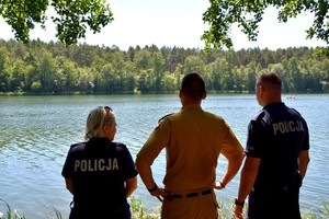 Policjanci i strażak odwróceni tyłem obserwują akwen wodny.