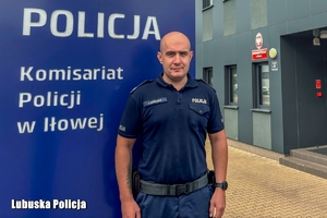 Umundurowany policjant pozuje do zdjęcia przy ściance z napisem: Policja, Komisariat Policji w Iłowej, w tle widoczny jest budynek komisariatu Policji.