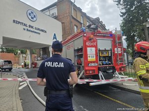 Wóz strażacki stojący przy szpitali, za taśmą stoi policjant i strażak.