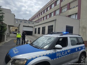 Radiowóz przed szpitalem na pierwszym planie, dalej dwaj policjanci stojący tyłem.
