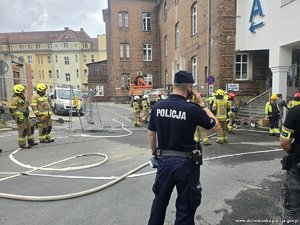 Policjant dzwoni telefonem komórkowym, wokół funkcjonariusze straży pożarnej.