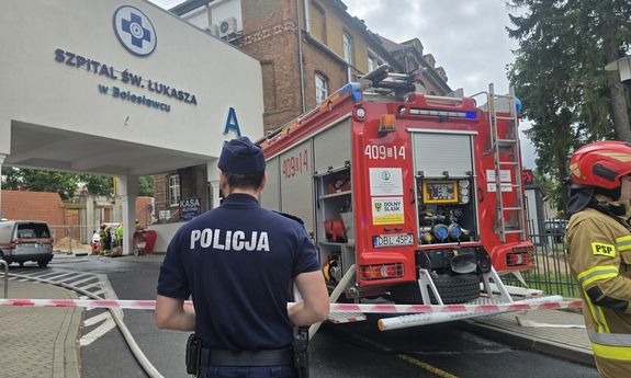 Wóz strażacki stojący przy szpitali, za taśmą stoi policjant i strażak.