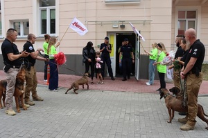Przewodnicy z psami stoją przed wejściem do szpitala, przed którym stoi dziewczynka z kobietą.