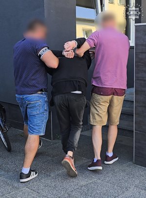 Na zdjęciu widoczni nieumundurowani policjanci, którzy prowadzą zatrzymanego mężczyznę.