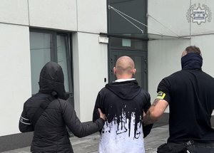 Nieumundurowani policjanci z opaskami z napisem Policja prowadzą zatrzymanego mężczyznę do budynku komendy.