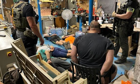 W magazynie na podłodze leżą zatrzymani, nad nimi stoją policjanci.