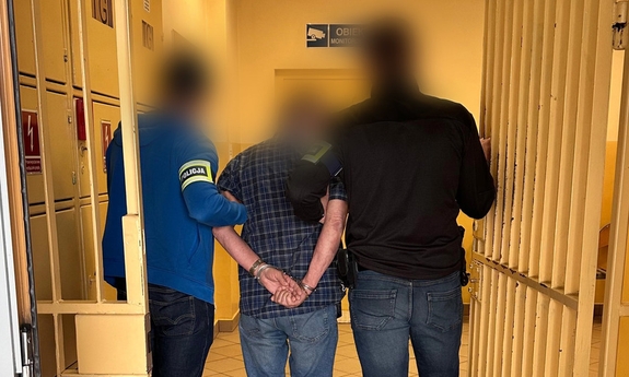 Mężczyzna trzymany przez policjantów w pomieszczeniu dla osób zatrzymanych.