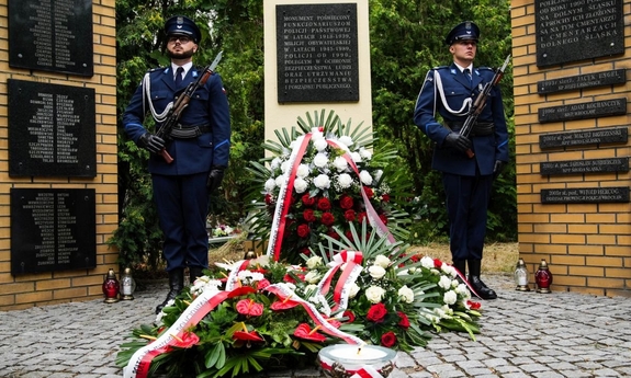 Dwaj policjanci stoją na warcie honorowej przy Tablicy Pamięci, pod którą  leżą złożone wieńce.
