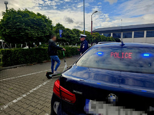 Na pierwszym planie tylna część radiowozu policyjnego, obok policjant i młody chłopak z hulajnogą.